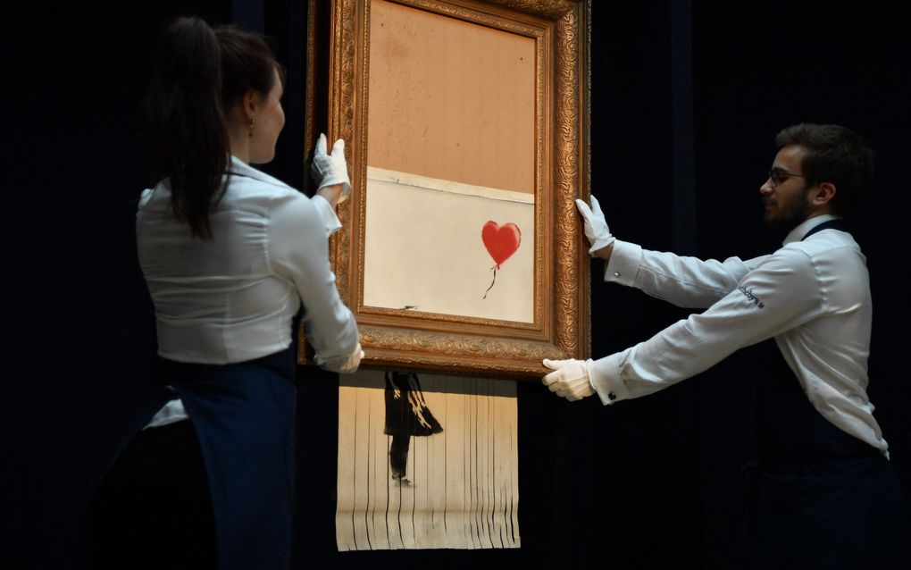 Twee mensen in witte blouses poseren met een kunstwerk dat half uit een lijst hangt en aan de onderkant is gerafeld.