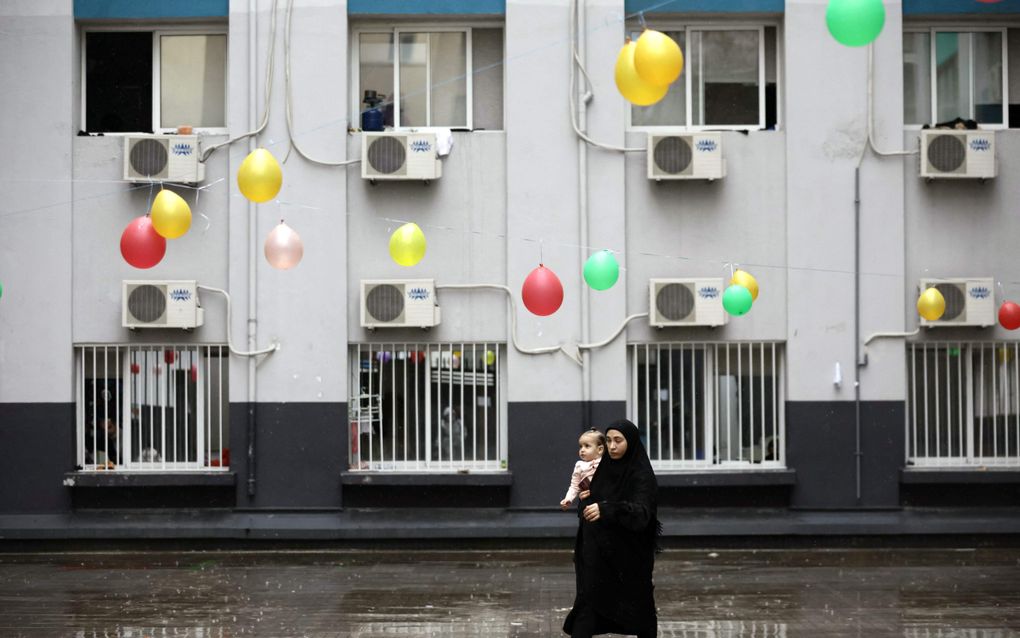 Vrouw in het zwart gekleed en met hoofddoek met een kind op de arm. Achter haar een wit gebouw. Gekleurde balonnen hangen op het plein. 
