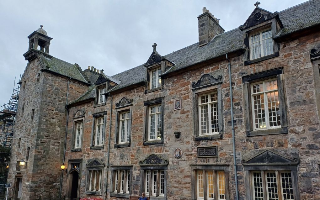 Een oud gebouw met heel veel kleine raampjes erin. Ernaast een grasveld met daarlangs roze en witte bloemen. Op een bankje zit een vrouw.