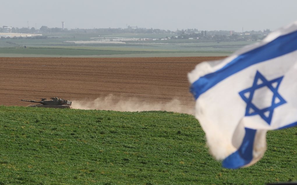Een tank rijdt door een vlak landschap. Op de voorgrond wappert een Israëlische vlag.