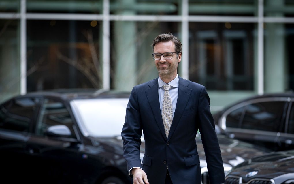 Een man met baard in pak met op de achtergrond een aantal donkere auto’s.