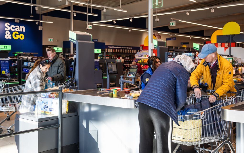 En man en een vrouw legen hun boodschappenkarretje bij een kassa in een supermarkt. Op de achtergrond rekent een andere vrouw haar boodschappen af bij een zelfscankassa.