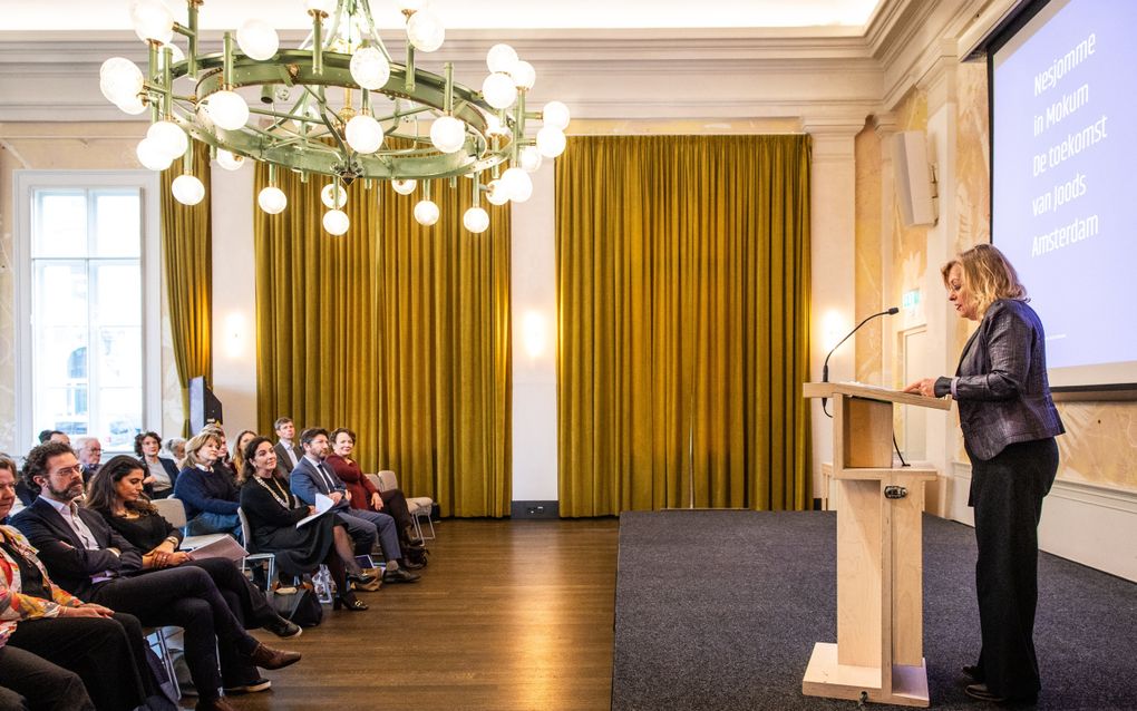 Een vrouw achter een katheder spreekt haar gehoor toe. Onder de aanwezigen de Amsterdamse burgemeester Femke Halsema.