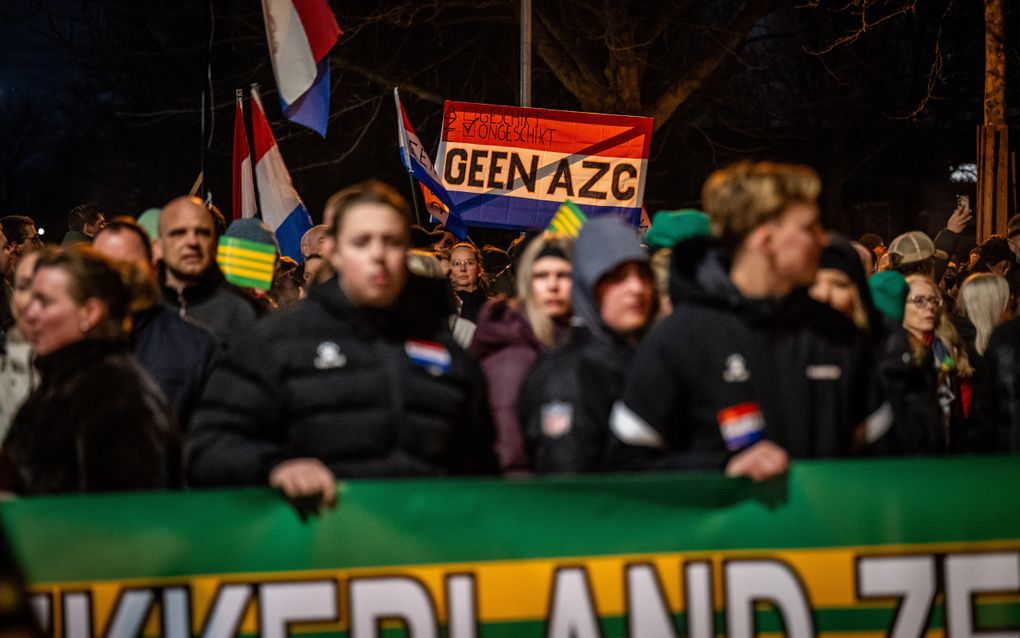 Tientallen mensen staan in de avond bij elkaar. Iemand houdt een Nederlandse vlag omhoog waarop staat: GEEN AZC.