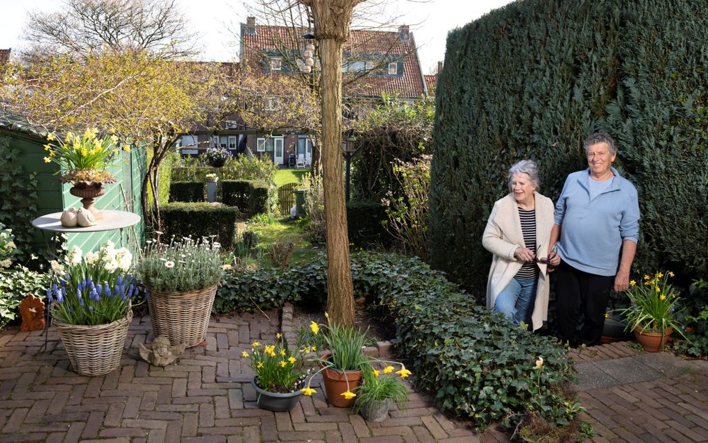 Een man en vrouw poseren naast elkaar in een tuin vol met hegjes, gras, terrasjes en potten met voorjaarsbloeiers. Geel, blauw, wit.