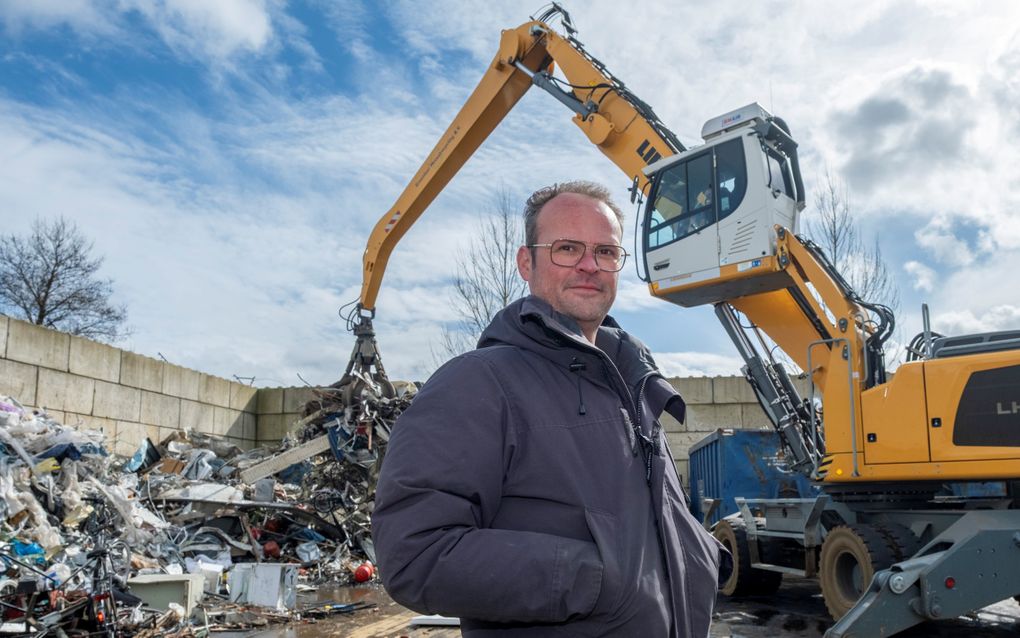 Man met bril staat met zijn handen in de zakken van zijn donkerblauwe jas voor een hoop schroot. Een gele kraan met cabine op hoogte staat achter de man. 