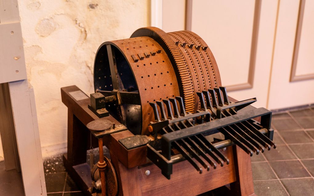 In een ruimte staat een roestbruine ronde trommel met tientallen gaatje. Aan de voorkant van de trommel zitten tien stokken die naar voren steken.