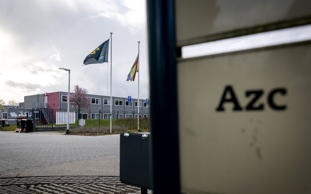 Op de voorgrond een bord waarop ”AZC” staat, op de achtergrond een woningencomplex met enkele vlaggenmasten ervoor.