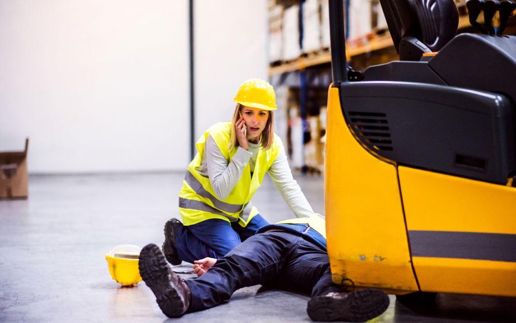 Een ongeluk in een magazijn. Een vrouw met een smartphone en haar collega liggen op de grond naast een heftruck.