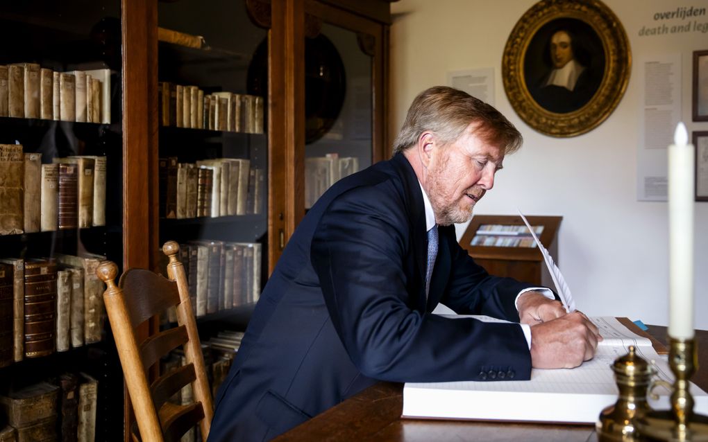 Een man met een baard, in donker pak, schrijft met een ganzenveer zijn naam in een gastenboek. Achter hem een boekenkast.