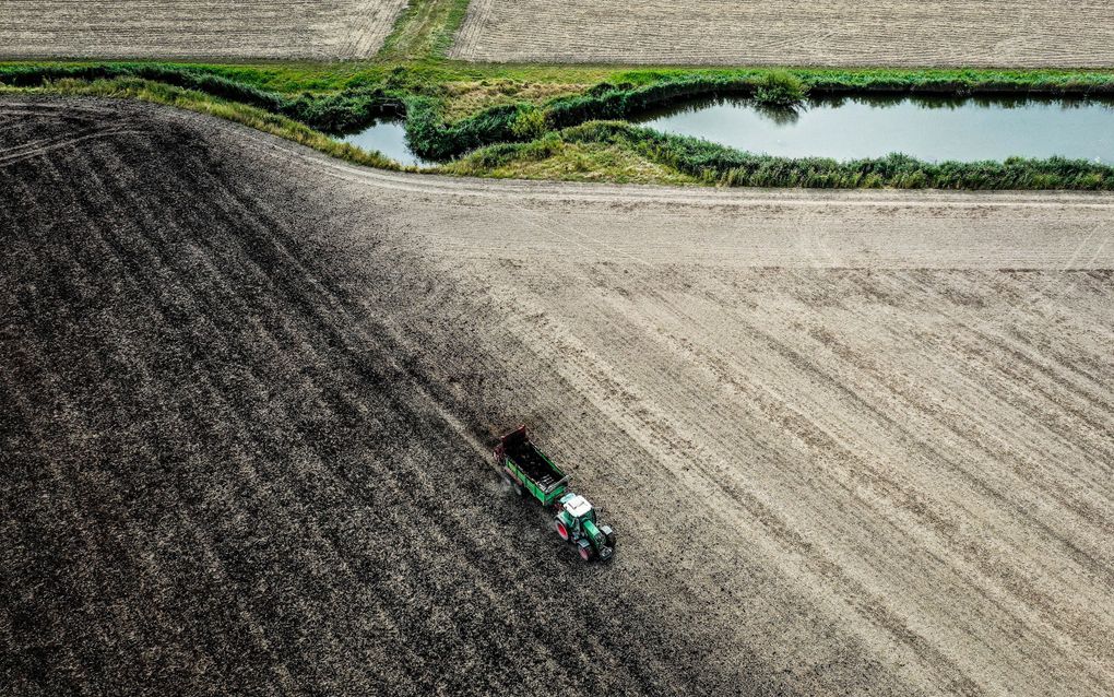 Luchtfoto van een trekker met aanhanger die mest uitrijdt over een akker. Langs de akker loopt een sloot.