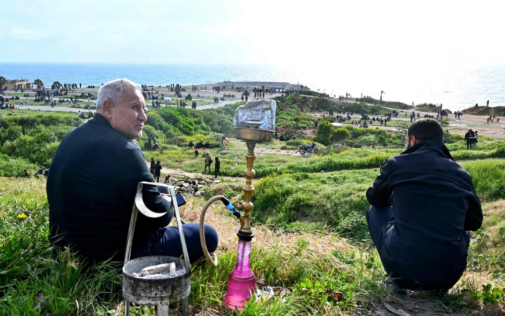 Twee mannen in donkere kleding zitten op een heuvel bij de zee. Tussen hen in een waterpijp