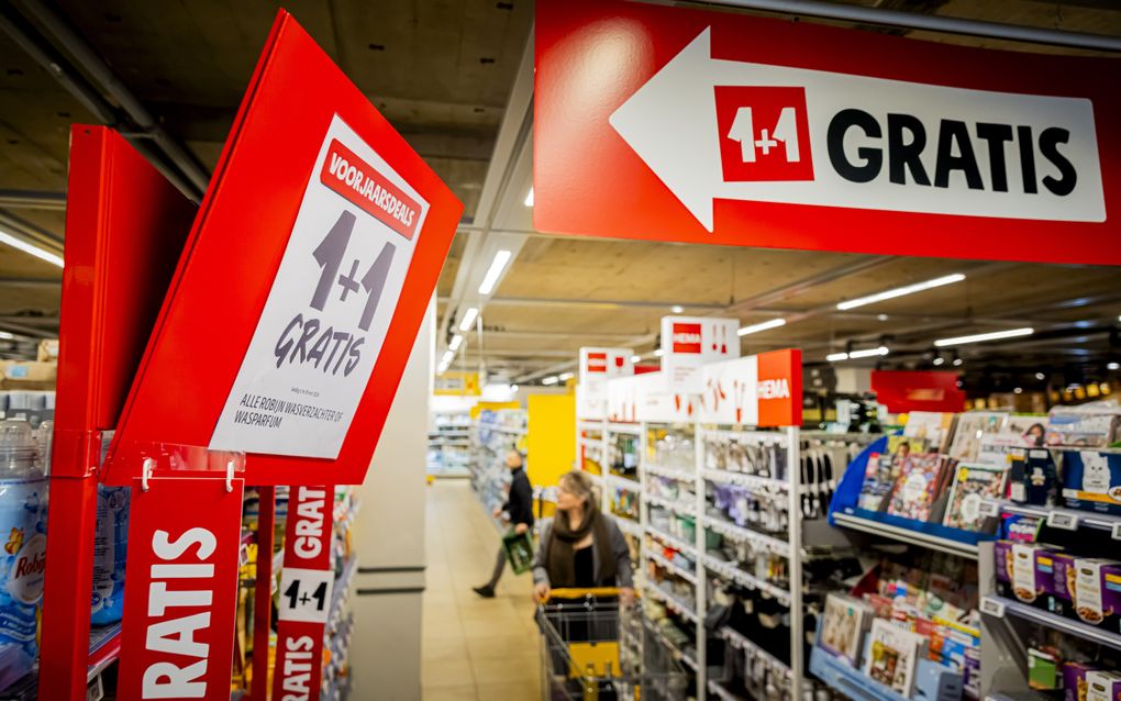 Een winkelende man en vrouw in een supermarkt, waarbij de vrouw een boodschappenkar voortduwt. Op een groot rood bord hangt een wit papier met daarop de aankondiging 1 plus 1 gratis.
