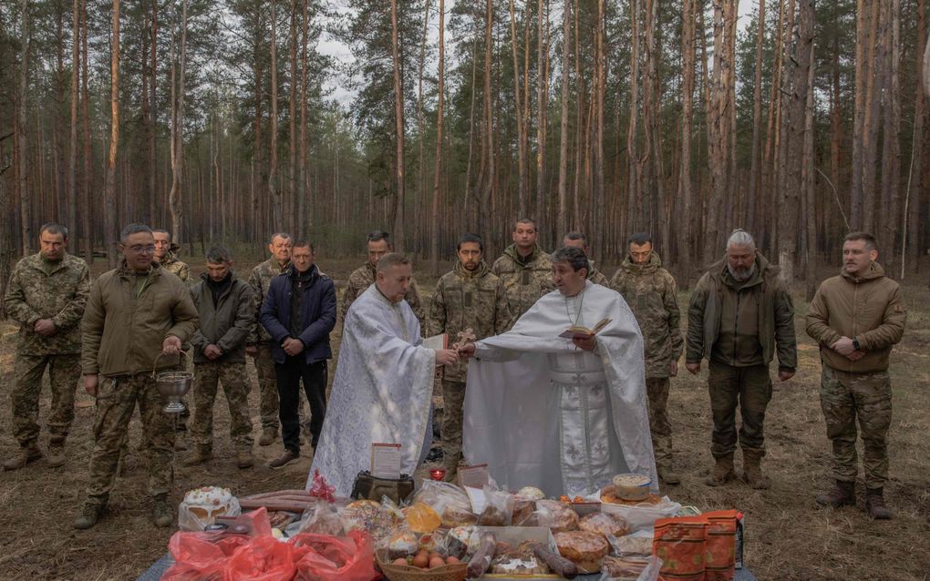 Mannen in witte gewaden voor een tafel met lekkers in een bos. Op de achtergrond militairen. 