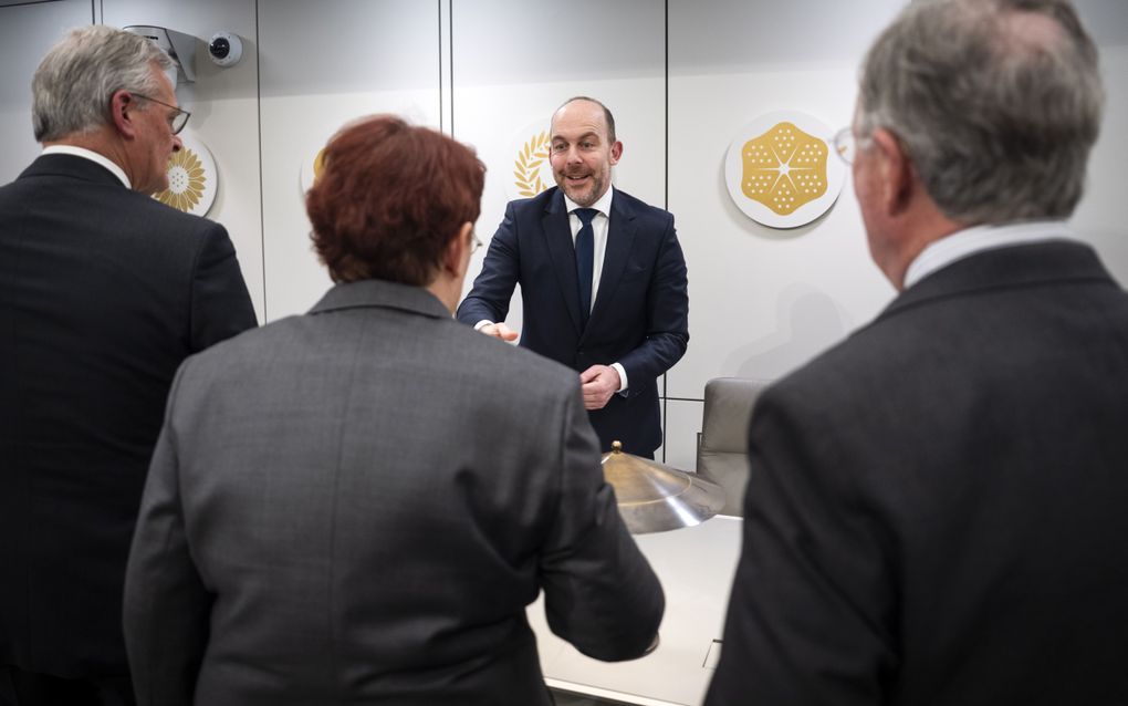 Een man in een donkerblauw pak schudt de handen van enkele mensen die voor hem staan. Deze mensen dragen donkere colberts.