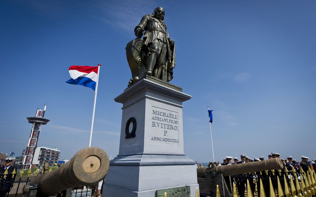 De Nederlandse vlag klappert naast een glimmend donker beeld op een blauwgrijze sokkel.
