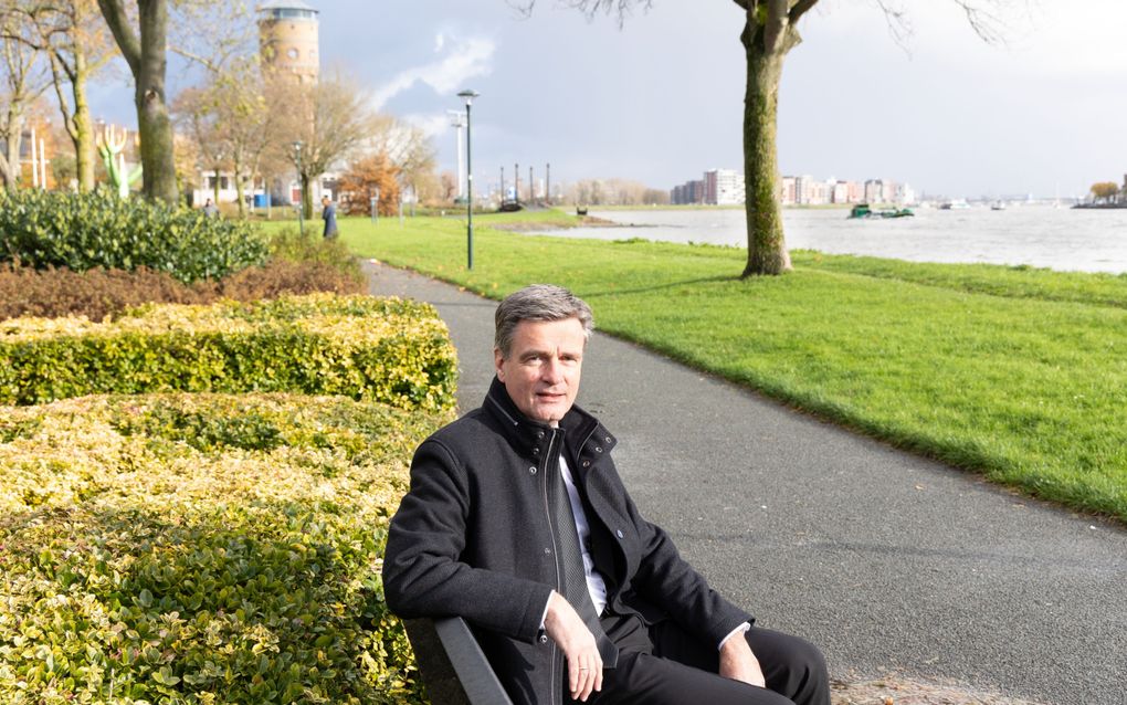 Een man in zwart pak, zittend op een bankje bij een rivier.