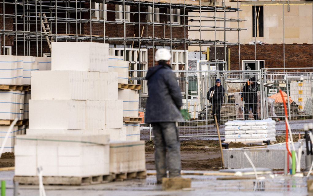 Een natte, modderige bouwplaats waar meerdere bouwvakkers in werkkleding en met helmen tussen stapels grote witte blokken en steigermaterialen werken aan de ruwbouw van een nieuw woongebouw.