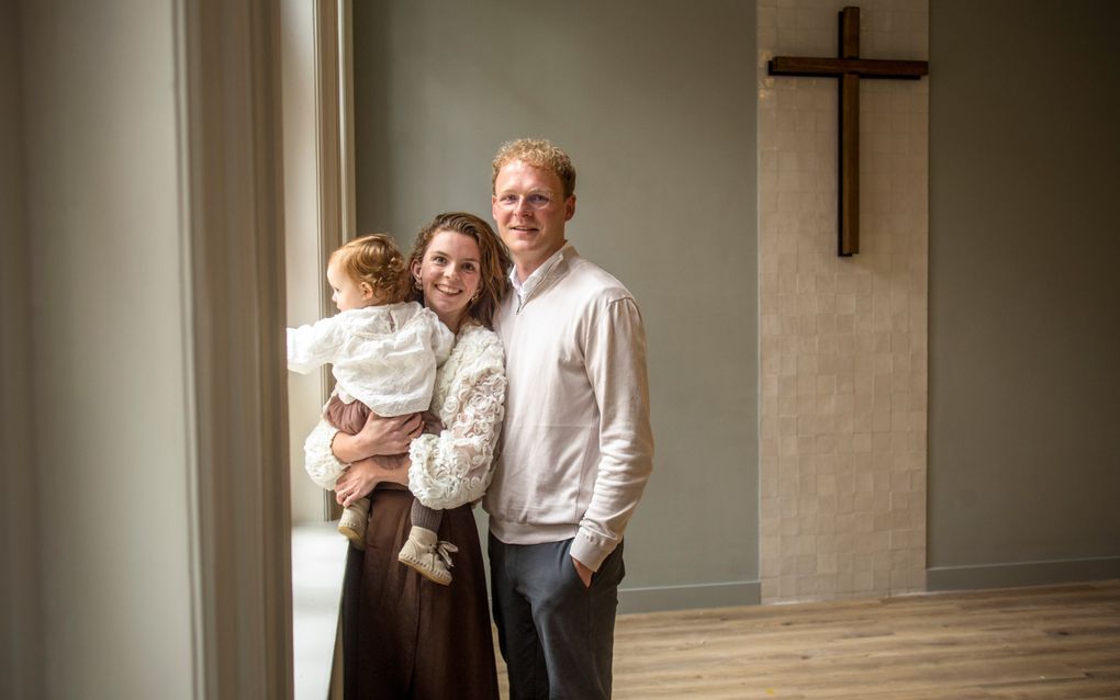 Een man met blond, krullend haar in een overhemd en beige trui, zijn vrouw met donkerblond, krullend haar met een klein meisje op de arm. Achter hen een muur met een betegelde strook met een kruis.