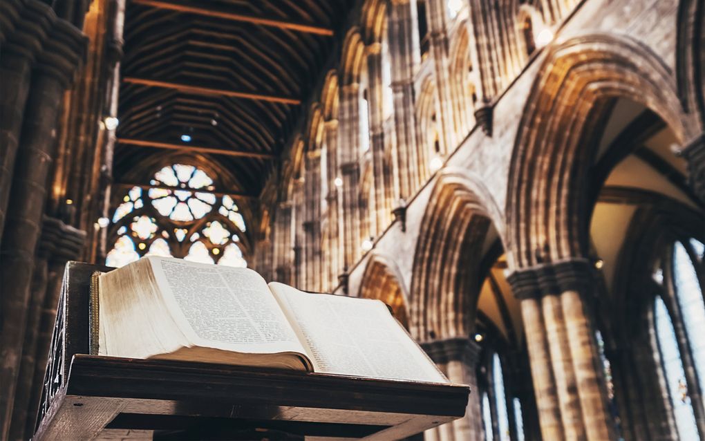 Een opengeslagen Bijbel op een oude houten katheder in het schip van een gotische kathedraal met glas-in-loodramen op de achtergrond.