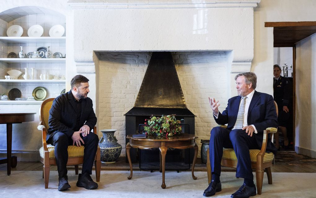 Een man in zwarte kleding leunt voorover terwijl hij naar de koning luistert.