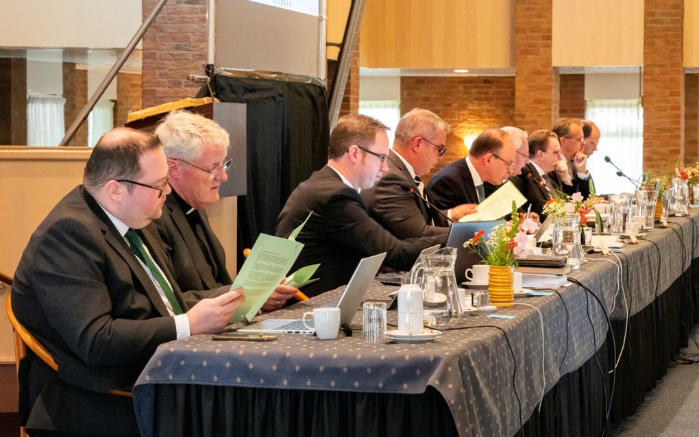 Lange vergadertafel, met daarachter negen mannen, gekleed in donkere pakken.