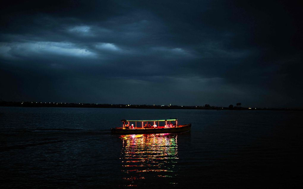 Een felverlicht bootje vaart op een donkere rivier. In de lucht hangen donkere wolken.