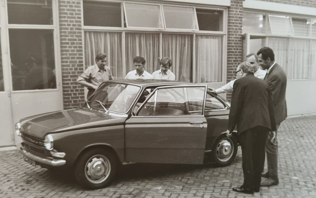 Foto in sepia van zes mannen die om een kleine auto heen staan en die bewonderen.  