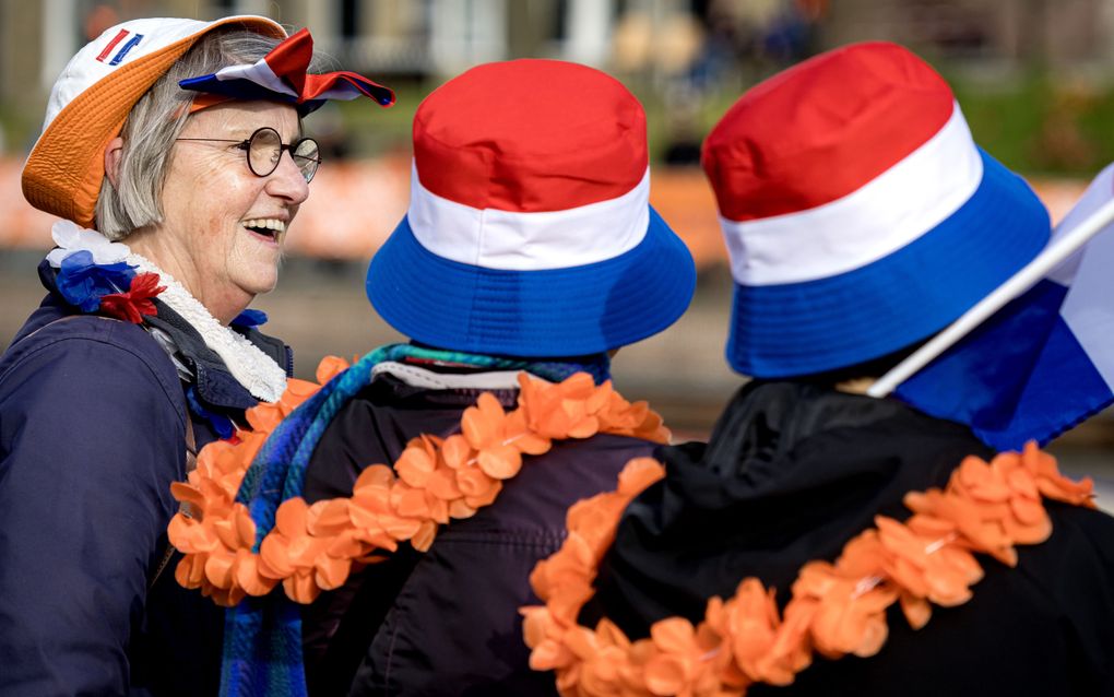 Een oudere vrouw en twee mensen (op de rug gezien) hebben rood-wit-blauwe hoedjes op en oranje en rood-wit-blauwe kettingen om.