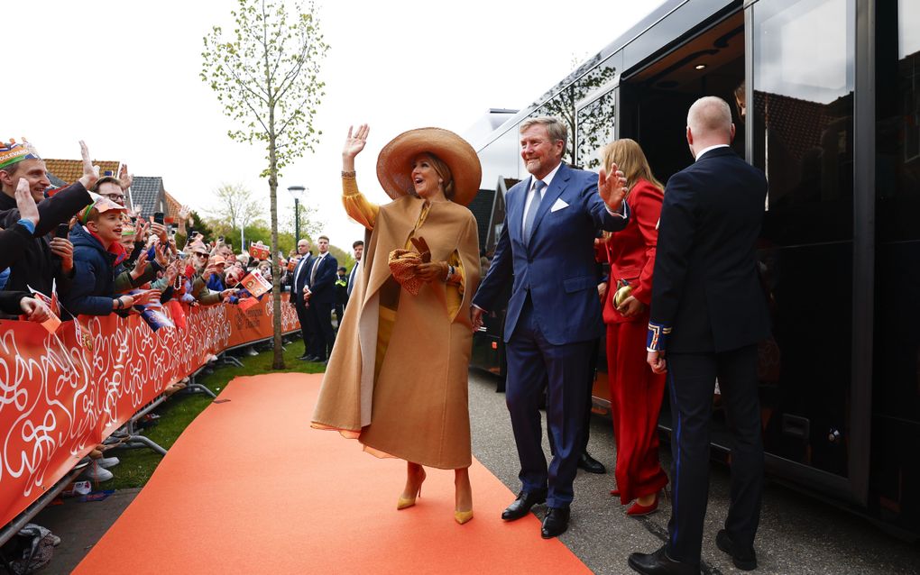 De koningin (gele jurk, jas, schoenen en hoed) en de koning (blauw pak en blauwe stropdas) stappen uit een zwarte bus en zwaaien naar mensen die achter dranghekken staan.