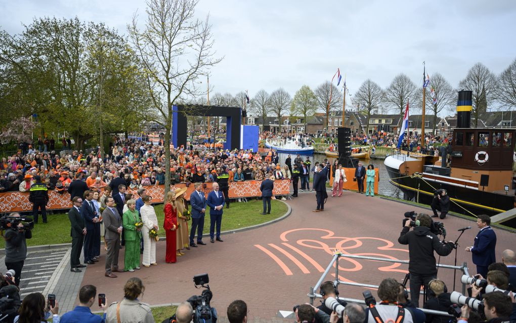 Rechts een man in blauw pak en met ketting achter een microfoon, links dertien leden van de koninklijke familie. Op de straat is het logo van Koningsdag 2026 in oranje getekend. Op de achtergrond veel publiek, op de voorgrond journalisten met camera’s.