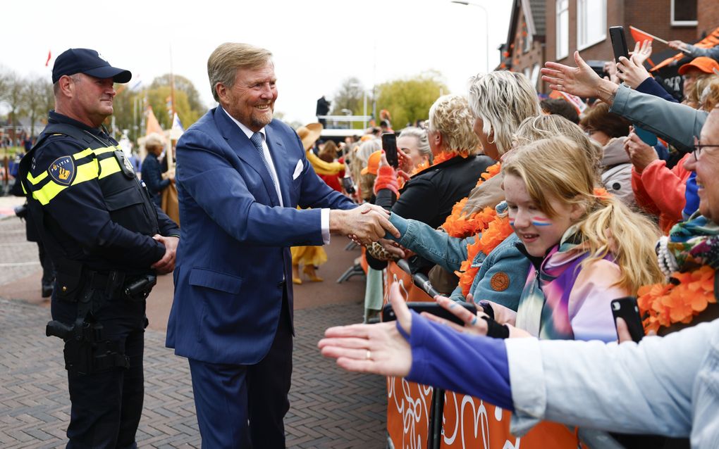 De koning, in blauw pak, schudt handen van mensen achter dranghekken, die oranje en rood-wit-blauw aanhebben. Achter hem staat een politieagent.