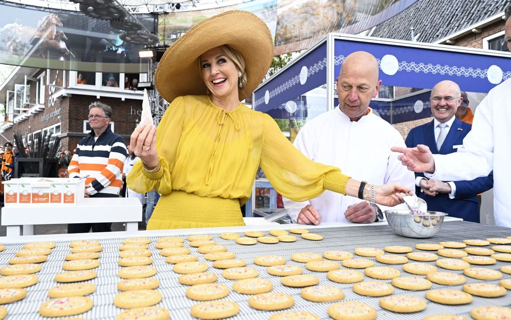 De koningin, in gele jurk en met gele hoed, houdt een klein spuitzakje omhoog en schept in een bakje met rood-wit-blauwe spikkels. Naast haar staat een bakker in een witte buis. Voor haar liggen koekjes. Rechtsachter staat een man in blauw pak.