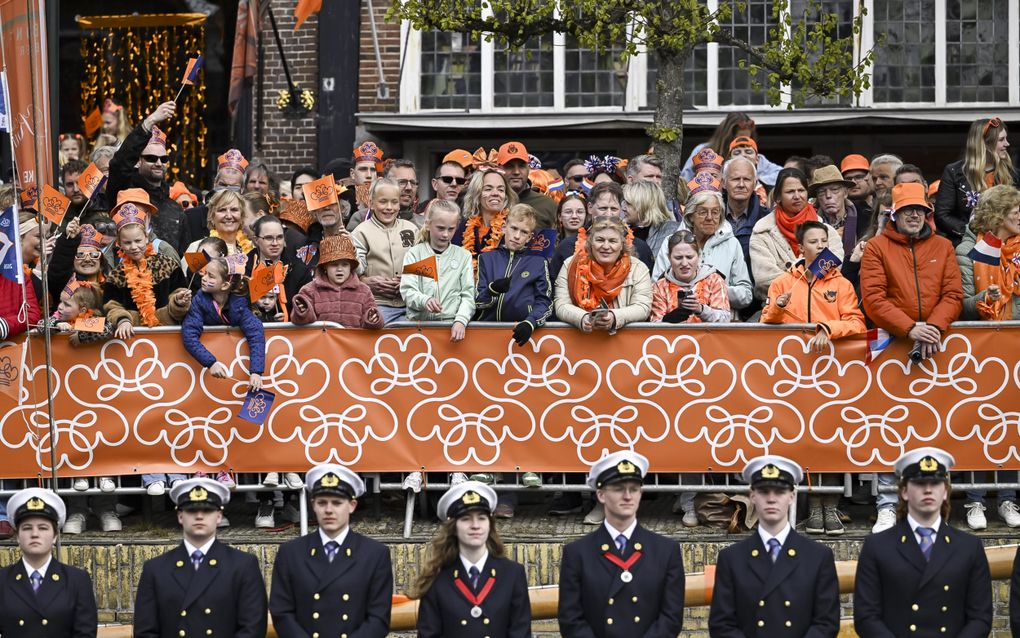 Mensen met oranje aan achter dranghekken, waarop een oranje doek is gespannen met witte lijnen die hartjes vormen. Op de voorgrond mensen met officieel uitziende blauwe pakken en witte petten.