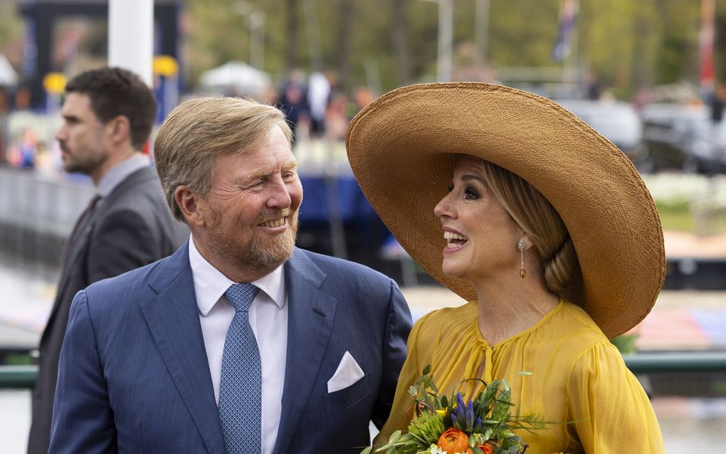 Links de koning, met blauw pak en blauw gestippelde das. Rechts de koningin, met gele jurk, hoed, oorbellen en tasje. In haar hand heeft ze een bosje bloemen. Ze lachen allebei.