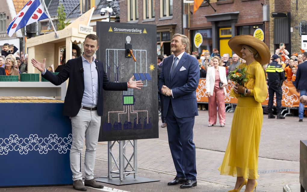 Links een man in colbert. In het midden een grote stroomschakelaar op een bord met het woord „stroomverdeling”. Rechts de koning en de koningin.