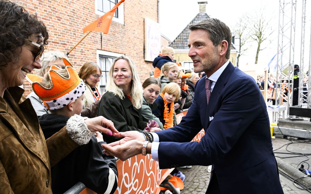 Een man met grijs haar, donkerblauw pak, paarsrode stropdas en verschillende armbanden loopt langs mensen achter dranghekken.