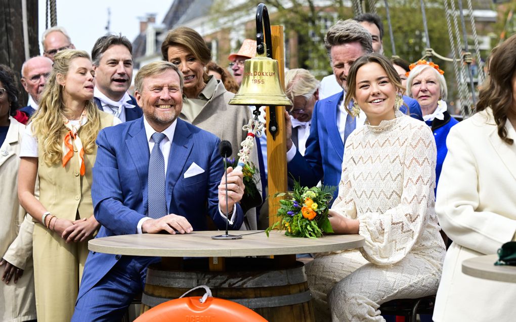 De koning zit aan een rond tafeltje en houdt het touw van een gouden scheepsbel vast, waarop staat: „Dokkum 1955”. Naast hem zit prinses Ariane, en achter hen staan verschillende mensen.