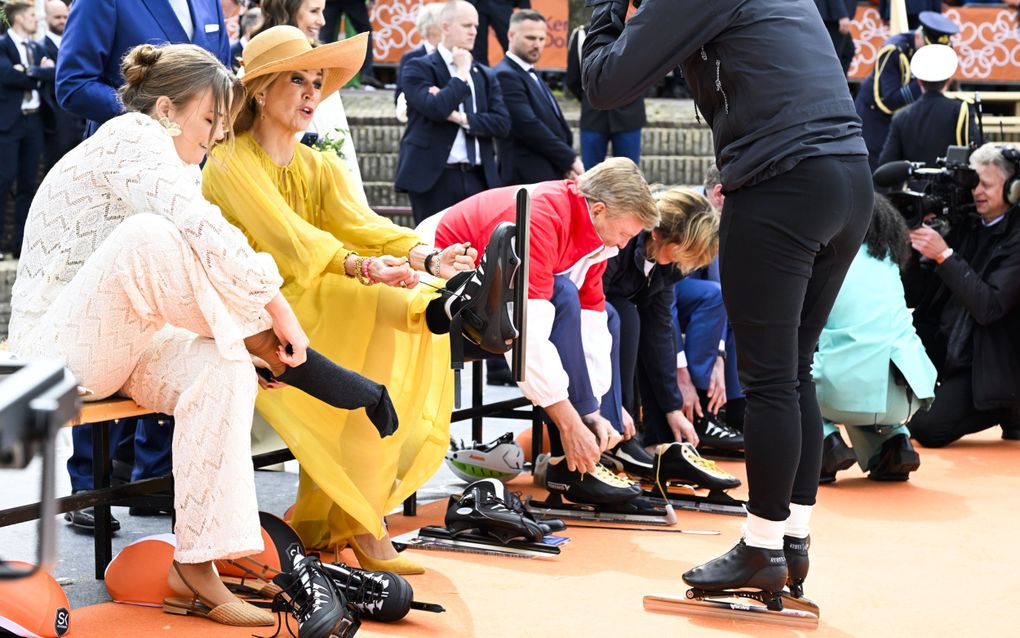 Links prinses Ariane, daarnaast koningin Máxima, daarnaast koning Willem-Alexander. Ze zitten op een bankje terwijl ze hun schaatsen aandoen. Voor hen staat een man in zwart, met schaatsen aan.