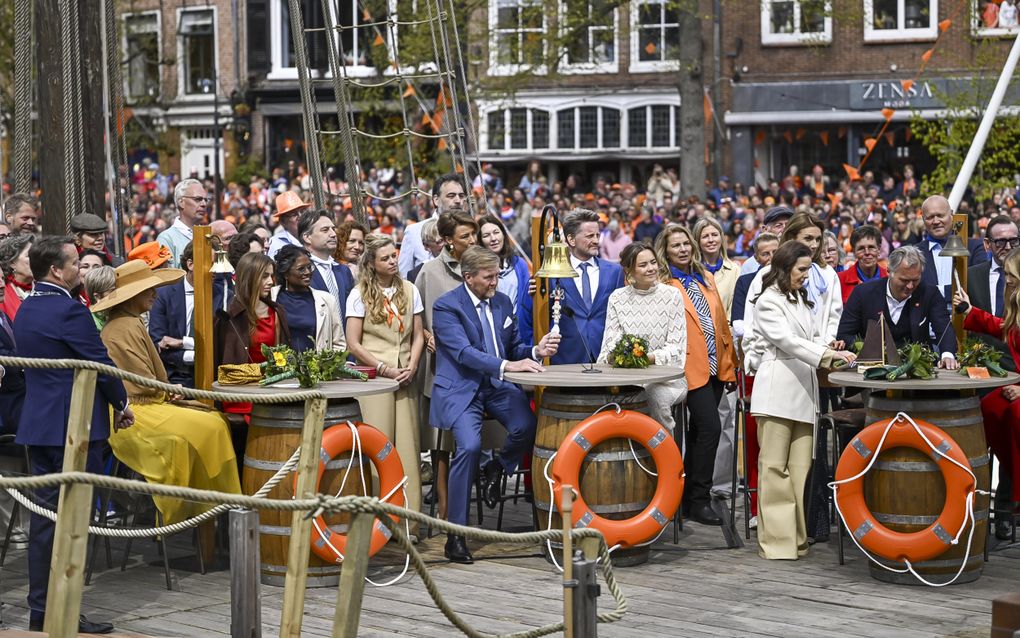 Drie ronde tafels staan op een schip. Bij elke tafel staat een scheepsbel. Bij iedere tafel staan en zitten leden van de koninklijke familie, en andere mensen.