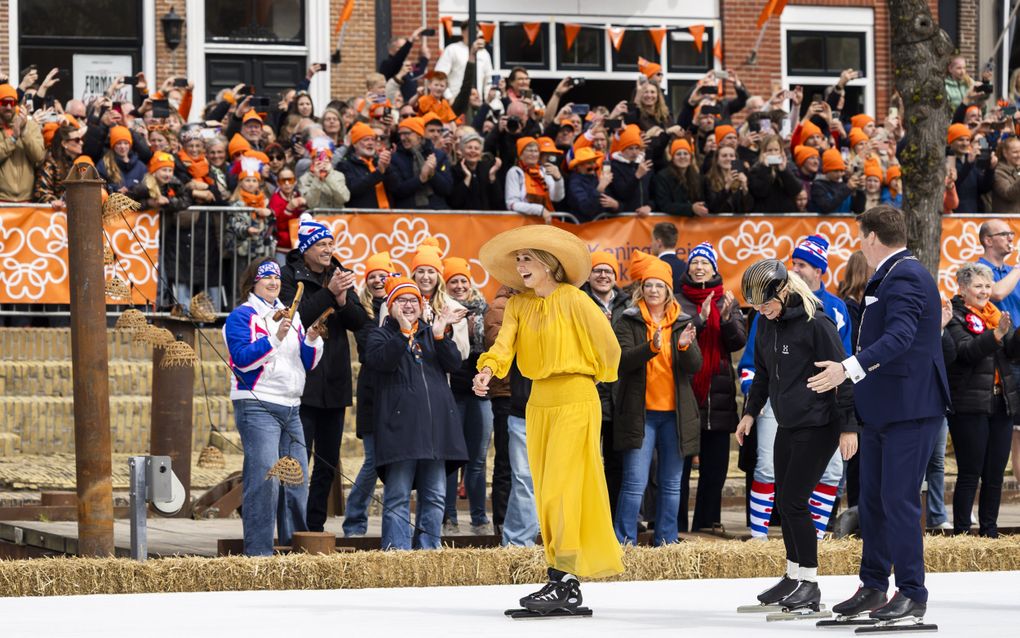 De koningin schaatst over wit kunstijs. Aan de rand staan hooibalen. Er staan mensen met oranje mutsen en mutsen met de Friese vlag te klappen aan de zijkant. Op de achtergrond staan mensen in oranje achter dranghekken.