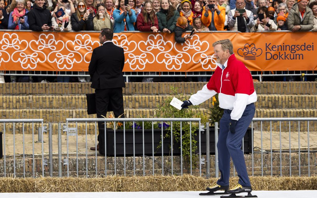 De koning, met blauwe broek en rood-witte trui, schaatst over kunstijs. Hij heeft een kaartje in zijn hand.