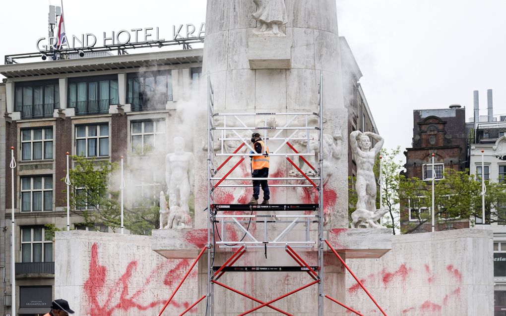 Mannen in oranje hesjes zijn met een hogedrukspuit in de weer voor een groot stenen monument waar rode verf op is gekalkt.