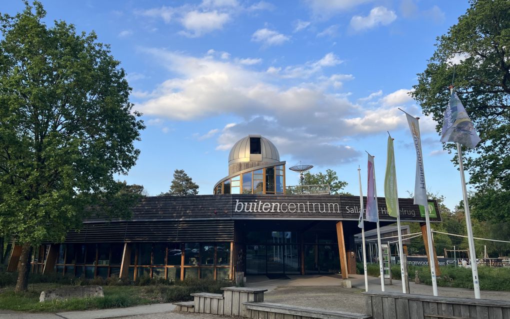 Laag houten gebouw met daarbovenop een toren met een zilverkleurige koepel. Links en rechts staan bomen met groen blad. In de lucht zijn enkele grijs-witte wolken te zien.
