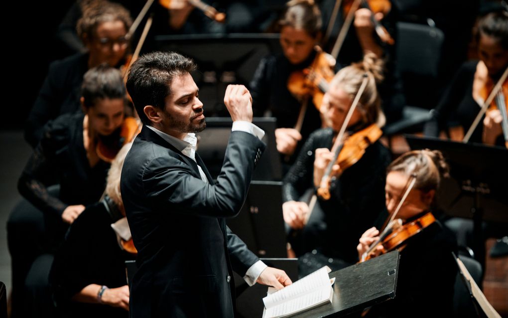 Een dirigent in een zwart pak staat met opgeheven rechterarm tussen de musici van een orkest.