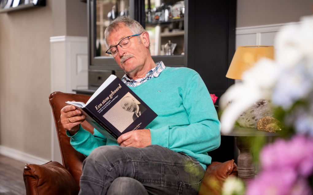 Een oudere man met een azuurblauwe trui in een bruine stoel; hij zit een boek te lezen.