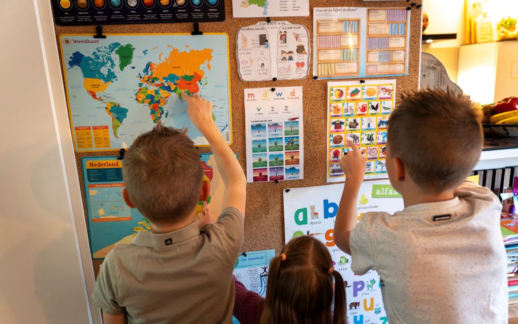 Drie kinderen staan voor een wand met allerlei leerzame posters, zoals een wereldkaart en platen met het alfabet.