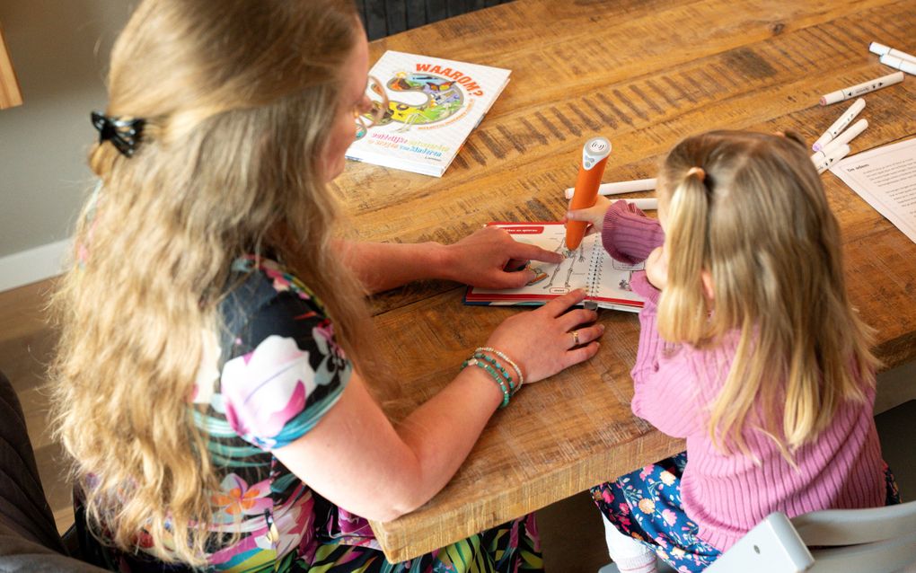 Een vrouw met lang blond haar en een meisje zitten samen aan tafel en lezen in een boekje.