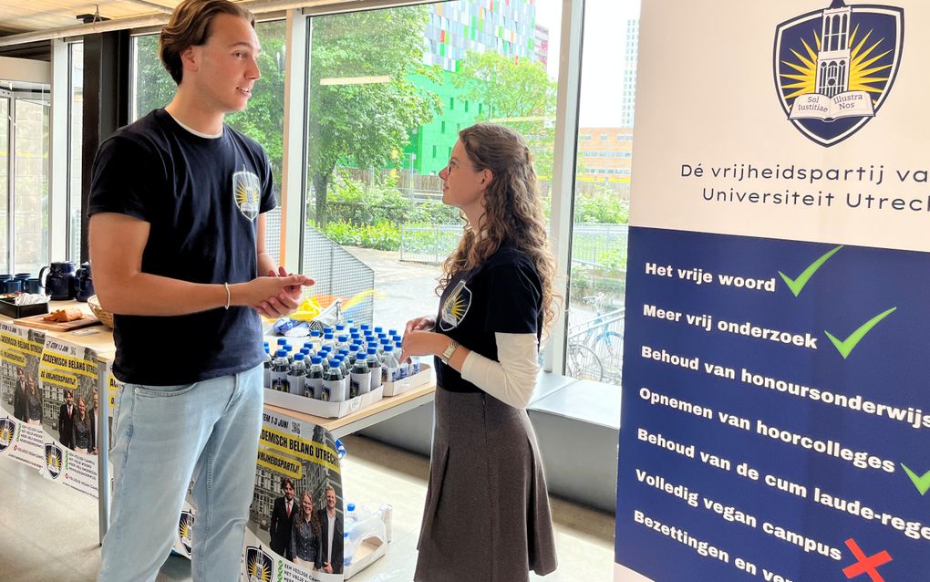 Een jongeman met bruin haar en een zwart shirt praat met een jonge vrouw in een zwart shirt en een grijze rok. 