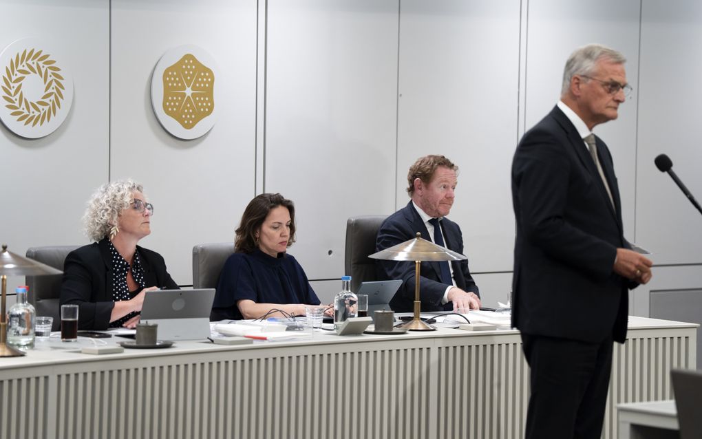Twee vrouwen en een man in pak zitten achter een bureau. Voor hen staat een man in pak achter een microfoon.
